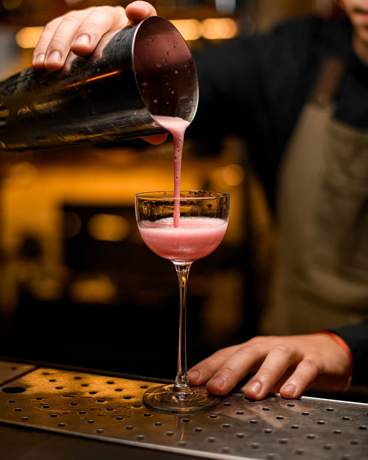 A bartender pouring a pink, foamy cocktail from a shaker into a coupe on the bar
